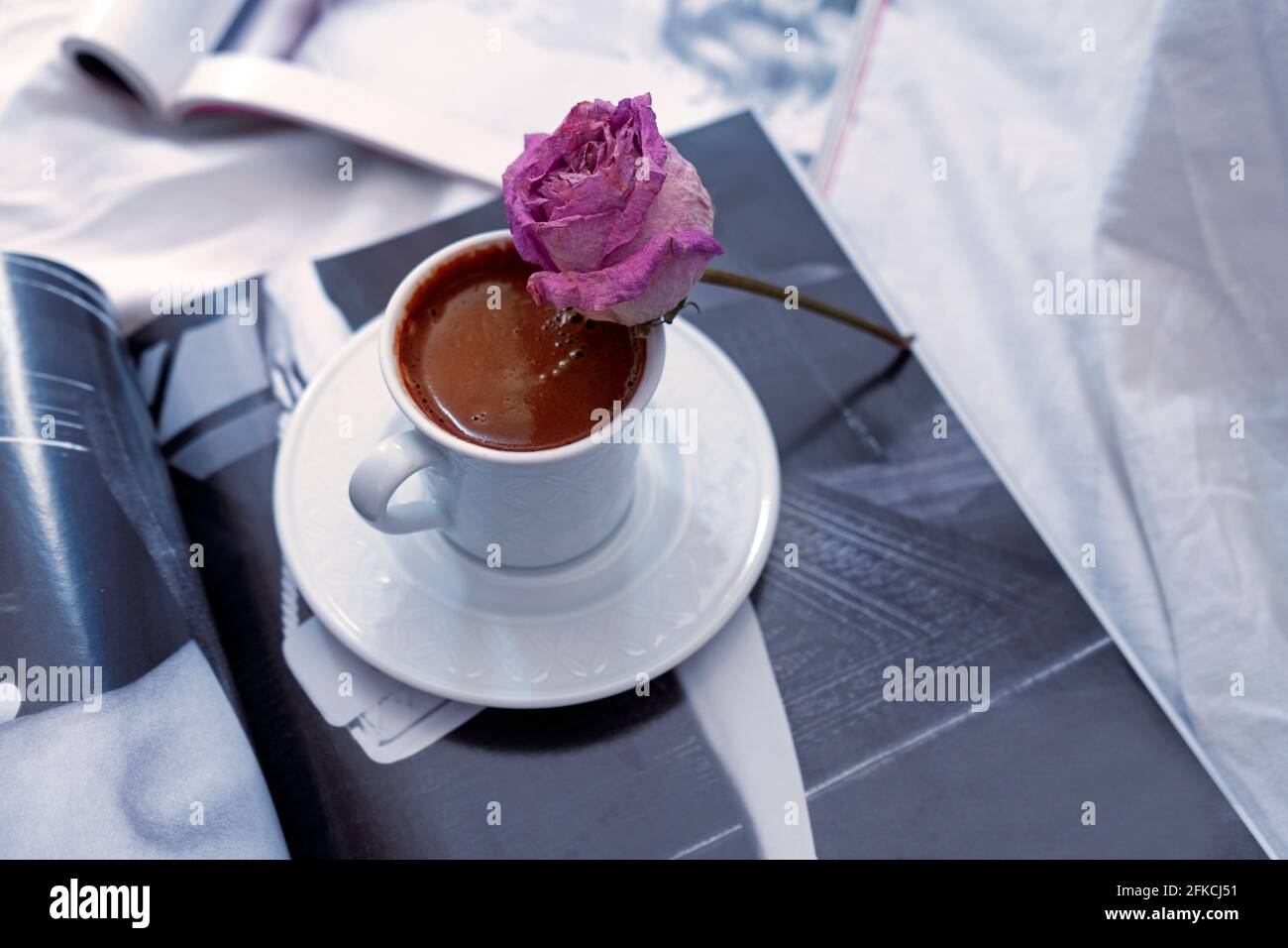 Ein türkischer Kaffeebecher und eine verdirte Rose auf dem Becher. Einige offene Zeitschriften darunter. Einige weiße Laken darunter. Romantische Morgenroutine. Stockfoto