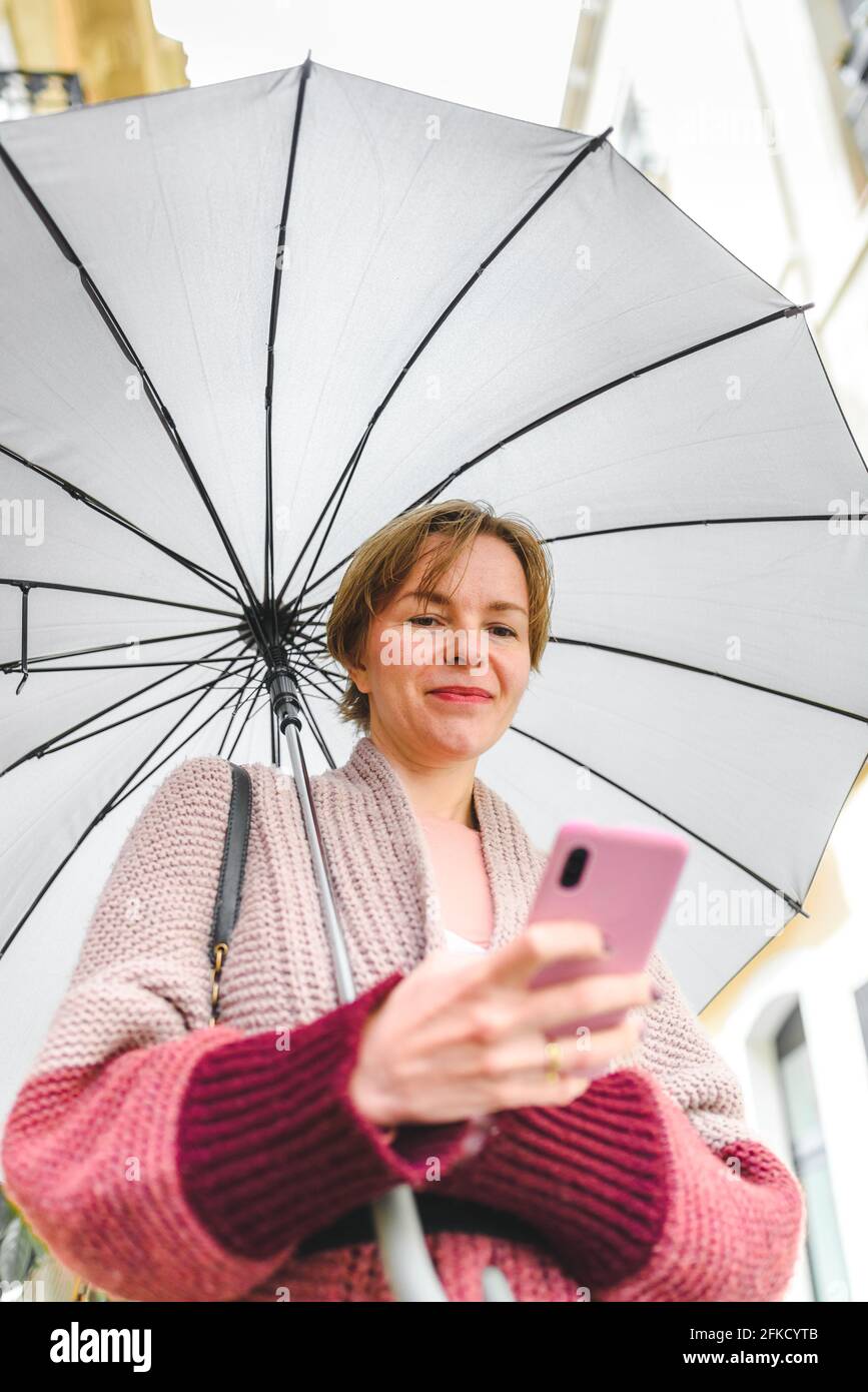 Vertikales Porträt einer blonden kaukasischen Frau, die ihr Telefon benutzt. Sie ist auf der Straße und hält einen Regenschirm Stockfoto