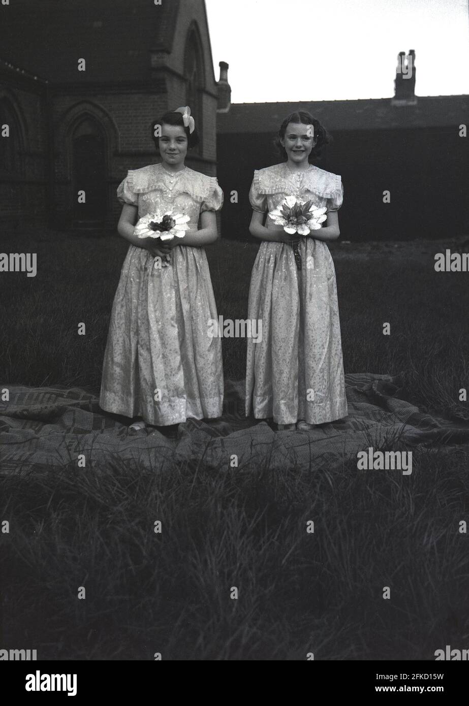 1958, historisch, draußen, auf dem Gelände einer Kirche, standen zwei Mädchen im Teenageralter in langen Kleidern und mit Blumensträußen auf einem Teppich für ein Foto vor dem Karneval der Königin im Mai. Sie würden die Maikönigin bei der Prozession begleiten, die entweder zu Fuß oder auf einem Festwagen während der Maifeiertage ritt, einer alten Tradition, die die Ankunft des Frühlings und des Sommers feierte. In vielen englischen Städten im Norden war die Parade mit den Sonntagsschulen der Church of England verbunden. Die Figur der May Queen und ihre Reinheit ist mit antiker Anbetung und Folklore verbunden. Stockfoto