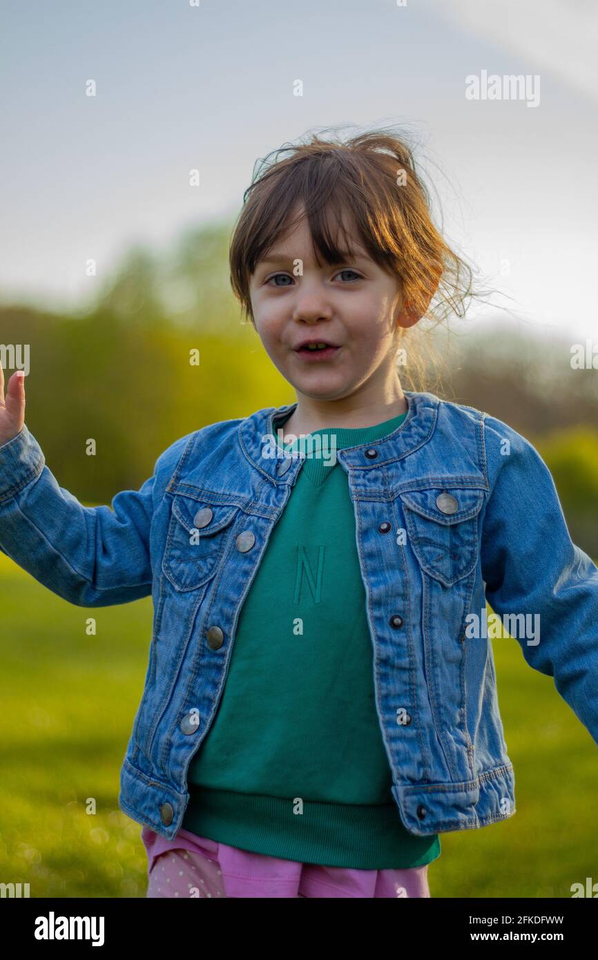 Porträt eines niedlichen, braunhaarigen, blauäugigen Mädchens, das an einem sonnigen Tag in einem Park läuft und eine blaue Jacke und einen grünen Pullover trägt Stockfoto