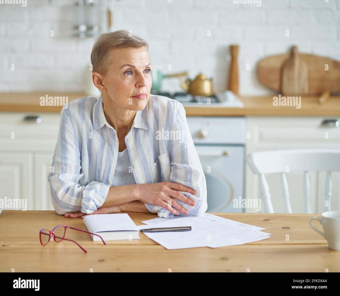 Schöne reife Frau, die beiseite schaut und an etwas denkt, während sie am Holztisch in der Küche sitzt und mit Dokumenten arbeitet. Senior People und Fernarbeitskonzept Stockfoto