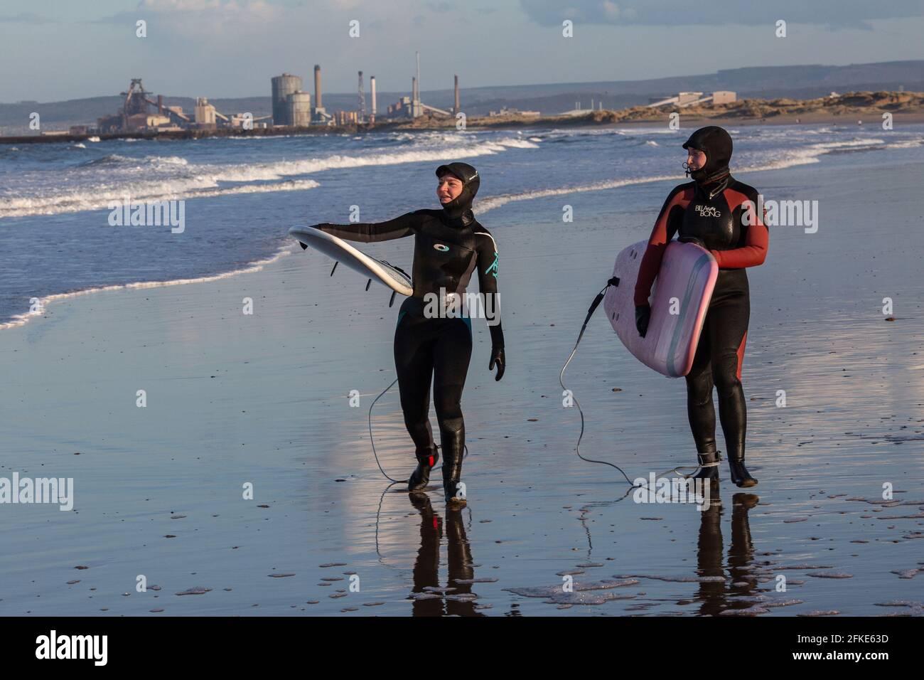 Olivia Harris (links) und ihre Freundin Rachel surfen in Seaton Carew, Nordostengland, mit Redcar Steelworks in der Ferne. Stockfoto