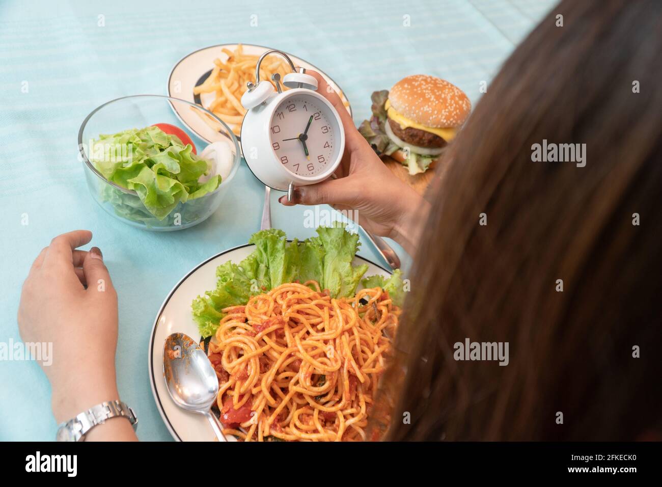 Die junge Frau hält die Uhr und ist bereit, einen Hamburger, Pommes Frites  und Spaghetti zum Frühstück zu essen. Konzept der Binge Eating Störung  (BETT) und entspannen Stockfotografie - Alamy