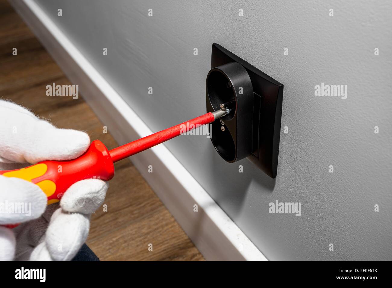 Ein Elektriker verwendet einen roten Schraubendreher, um eine schwarze, doppelt geerdete Steckdose in den Elektrokasten in der Raumwand zu Schrauben. Stockfoto