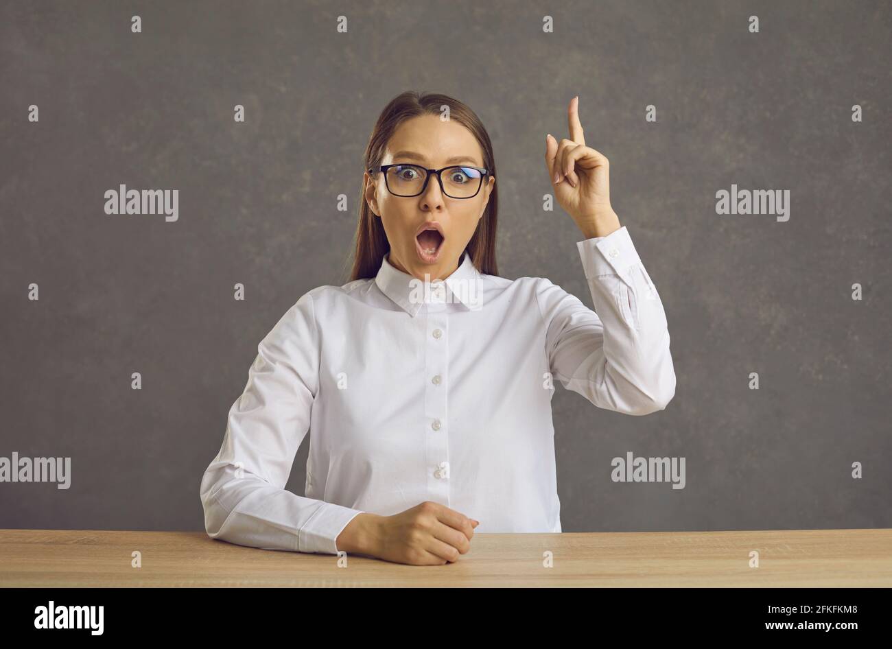 Frau in einer Brille sitzt an einem Schreibtisch und zeigt auf sie Finger auf überrascht von einer klugen Idee Stockfoto