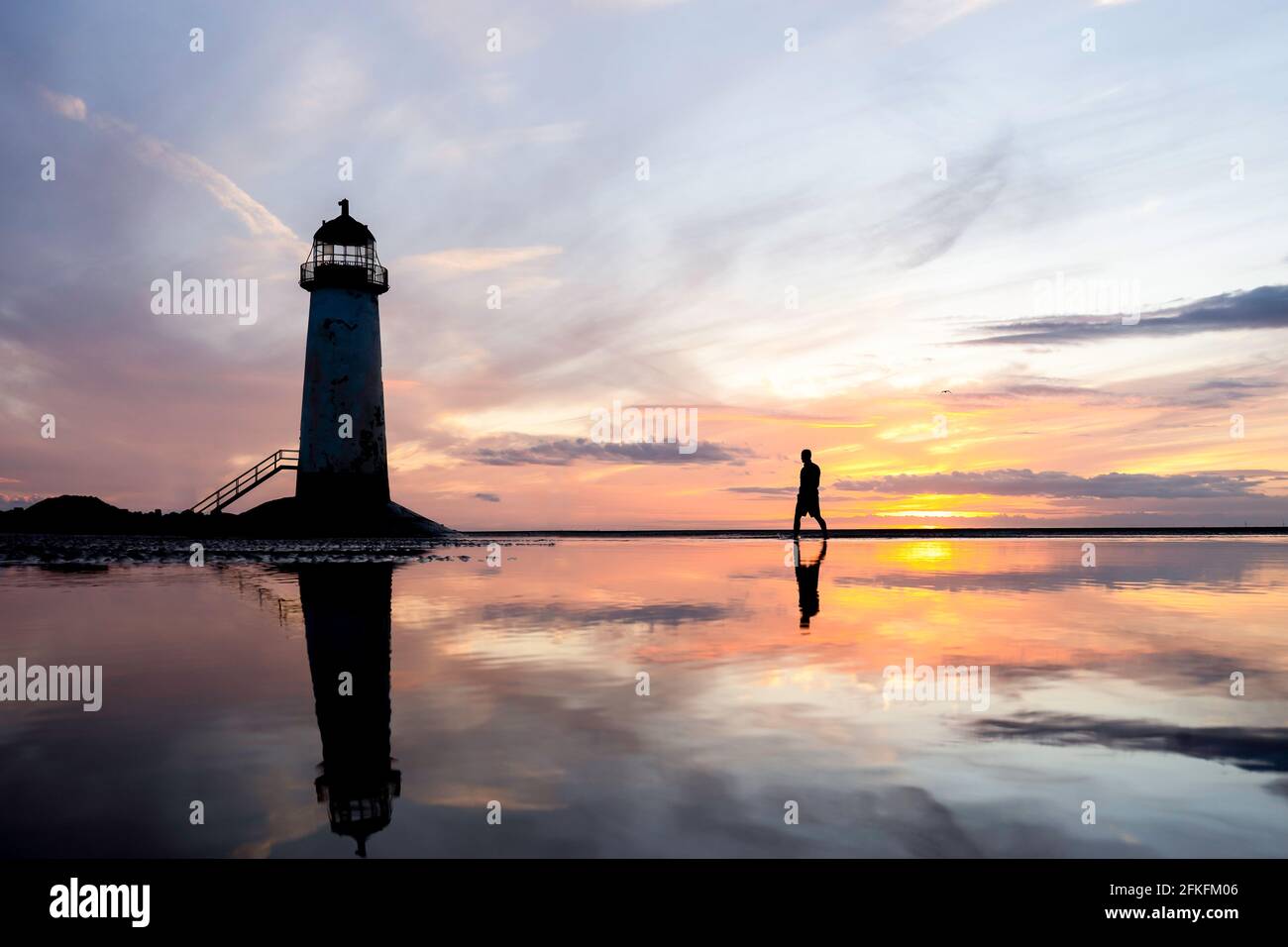 Leuchtturm steht im Wasserbecken atemberaubende Sonnenuntergangsreflexion im Meerwasser reflektiert. Einzelmann, der die Küste von Nordwales erkundet, Sand am Strand Stockfoto