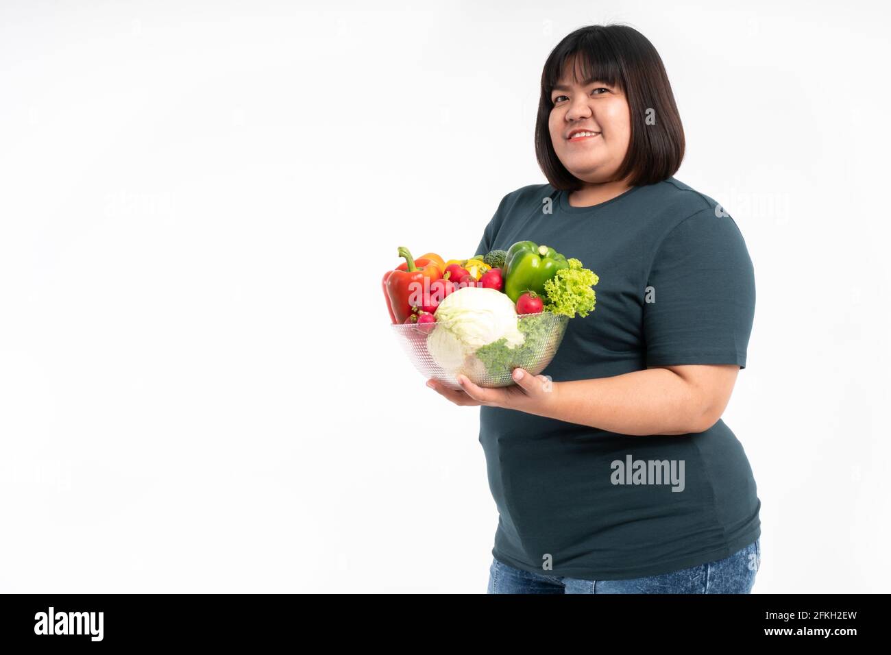 Glückliche übergewichtige asiatische Frau, die einen Gemüskorb hält und auf einem isolierten weißen Hintergrund lächelt, kommt das Konzept der guten Gesundheit vom Essen einer Nutritiou Stockfoto