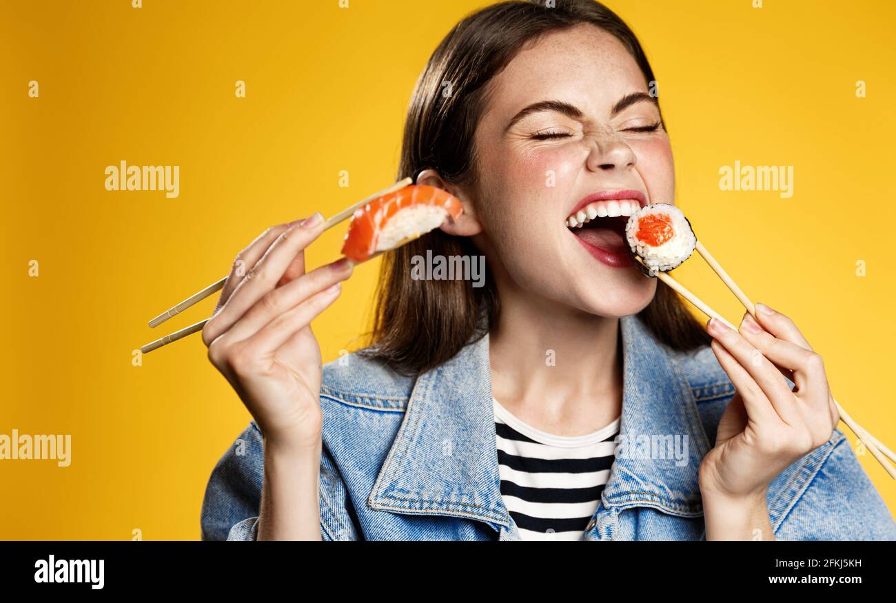 Glückliches Mädchen isst Sushi mit Zufriedenheit, hält Sashimi und beißt Sushi-Rolle mit Lächeln, bestellen köstliche Takeaway in Smartphone-App, zufrieden mit schnell Stockfoto