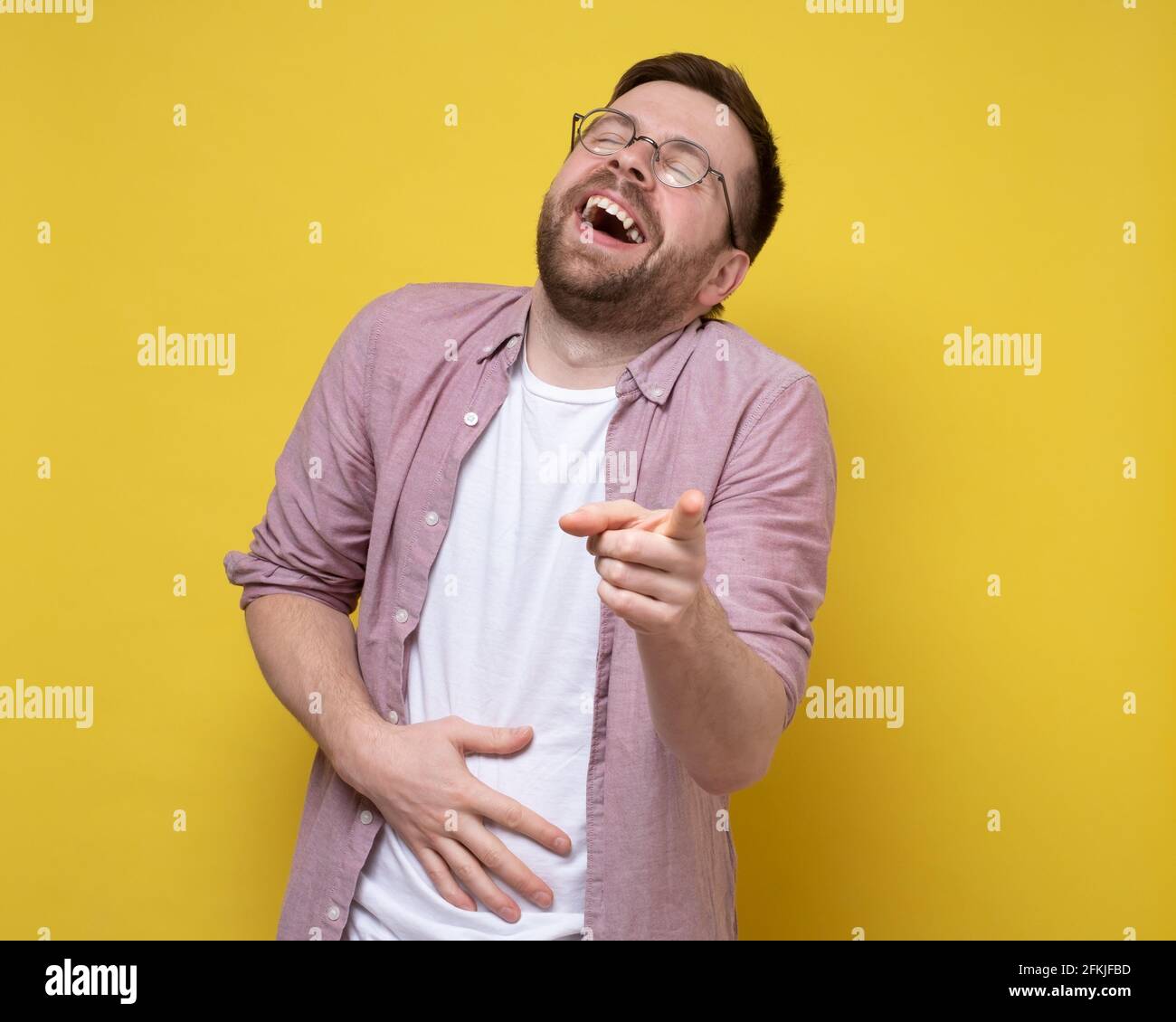 Mann in runden Brillen und legerer Kleidung lacht laut und spöttisch, hält den Bauch und zeigt mit dem Finger auf die Kamera. Stockfoto