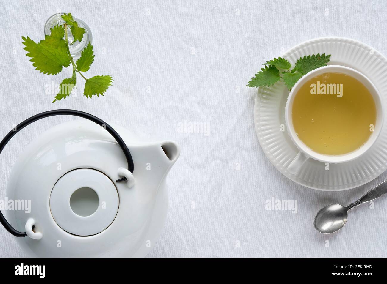 Tasse Brennnessel-Tee, frische Brennnesselkräuter. Flache Tischdecke, Draufsicht, aus weißer textiler Tischdecke. Weiße Teekannen aus Keramik. Stockfoto