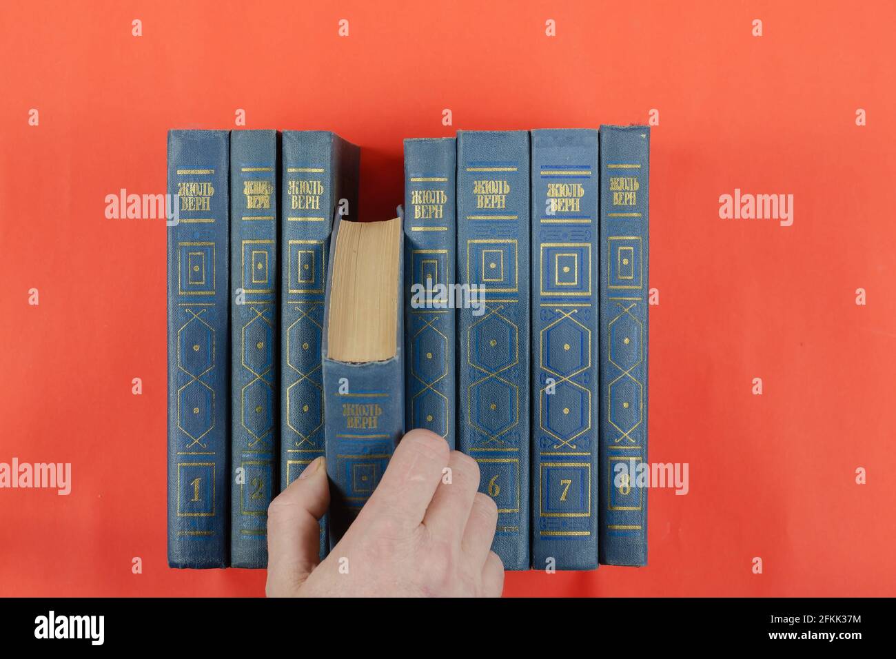 Bücher der klassischen Literatur auf rotem Hintergrund. Aus den gesammelten Werken von Jules Verne extrahiert eine Männerhand in 8 Bänden ein Buch. Hardcover-Bücher Stockfoto