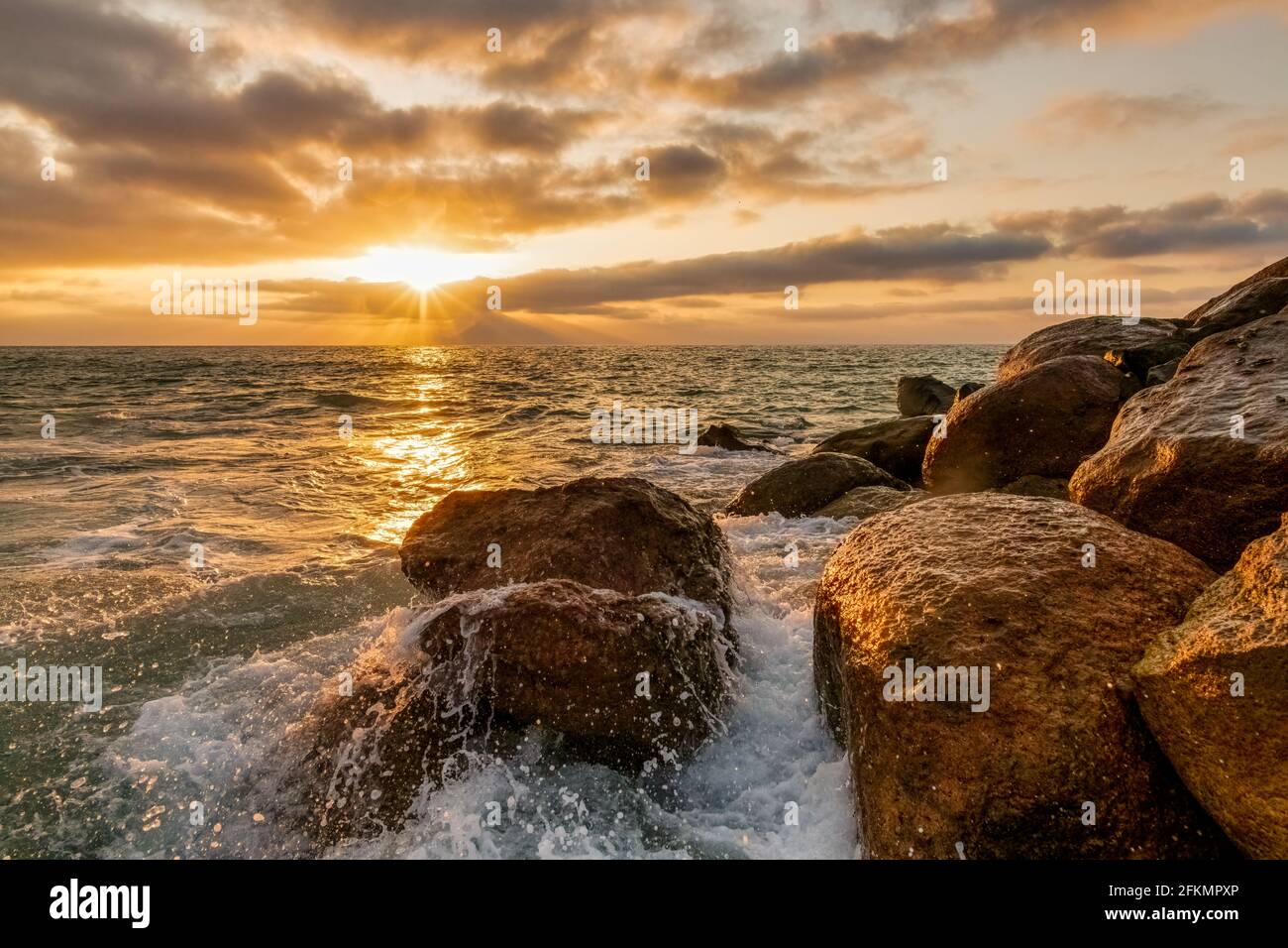 Eine Welle bricht und spritzt auf Sea Rocks AS Die Sonne untergeht am Meereshorizont Stockfoto