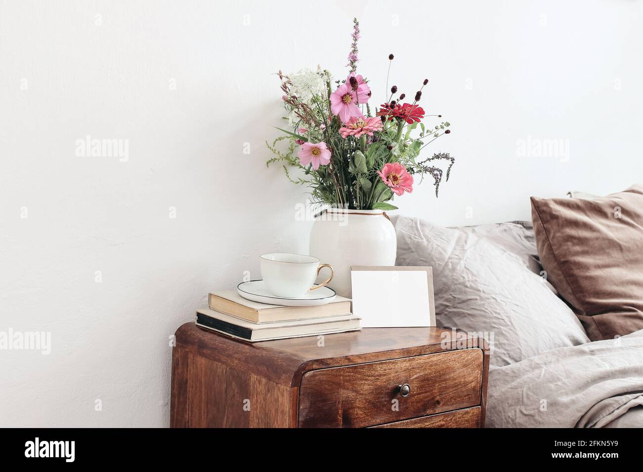Tasse Kaffee und Bücher auf dem Nachttisch aus Holz im Retro-Stil. Modell mit leerer Grußkarte. Weiße Keramikvase. Buquet von Kosmos und Zinnia Blumen. Beige Stockfoto