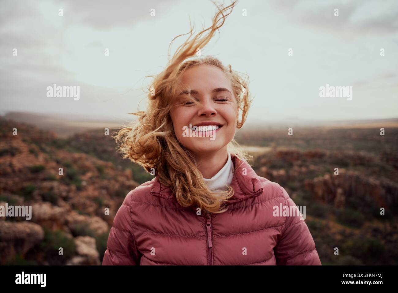 Lächelnde junge, selbstbewusste Frau mit geschlossenen Augen und frischem Wind Gegen das Gesicht, das auf einem Berghang steht Stockfoto