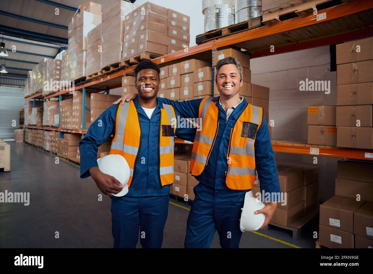 Lächelndes Porträt zweier ethnischer Lagerarbeiter, die in Sicherheit sind hardhat in der Hand im Werk Stockfoto