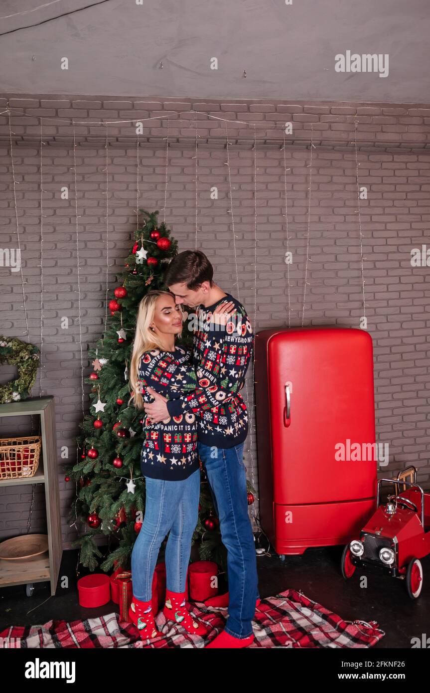 Ein Mann mit einem Mädchen in einem Pullover und Jeans Umarmen und küssen Sie auf dem Hintergrund eines Weihnachtsbaums Und rote Geschenke Stockfoto