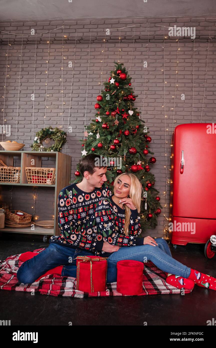 Ein Mann mit einem Mädchen in einem Pullover und Jeans Umarmen und küssen Sie auf dem Hintergrund eines Weihnachtsbaums Und rote Geschenke Stockfoto