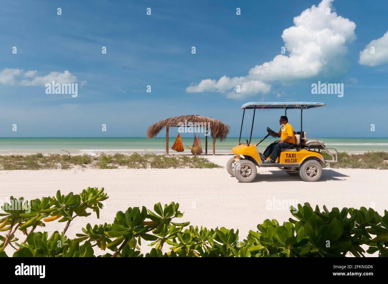 Ein gelbes Golfauto-Taxi an einem tropischen Strand auf Holbox Island in Mexiko. Im Hintergrund eine Hütte mit Hängematten und das Karibische Meer Stockfoto