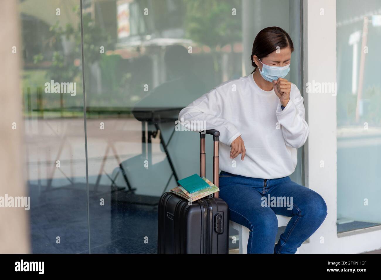 Reisen . Eine kranke Frau in einem weißen Langarm-Hemd und Jeans, trägt  eine Maske und hustet, sitzen auf dem Stuhl mit Koffer, Karte, Notizbuch  und Rucksack o Stockfotografie - Alamy