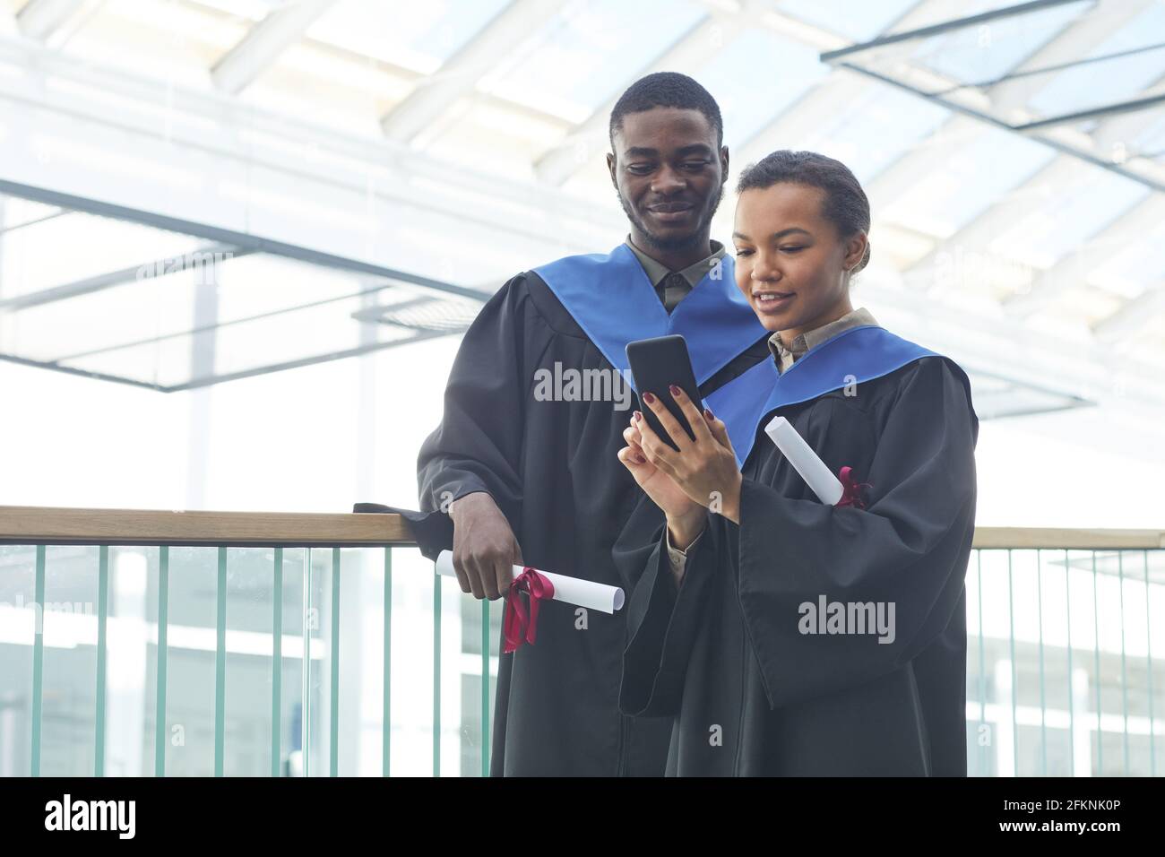 Waist-up-Porträt eines jungen afroamerikanischen Paares mit Abschlusskleidern, während es in einem modernen Universitätsinnenraum Selfie macht Stockfoto