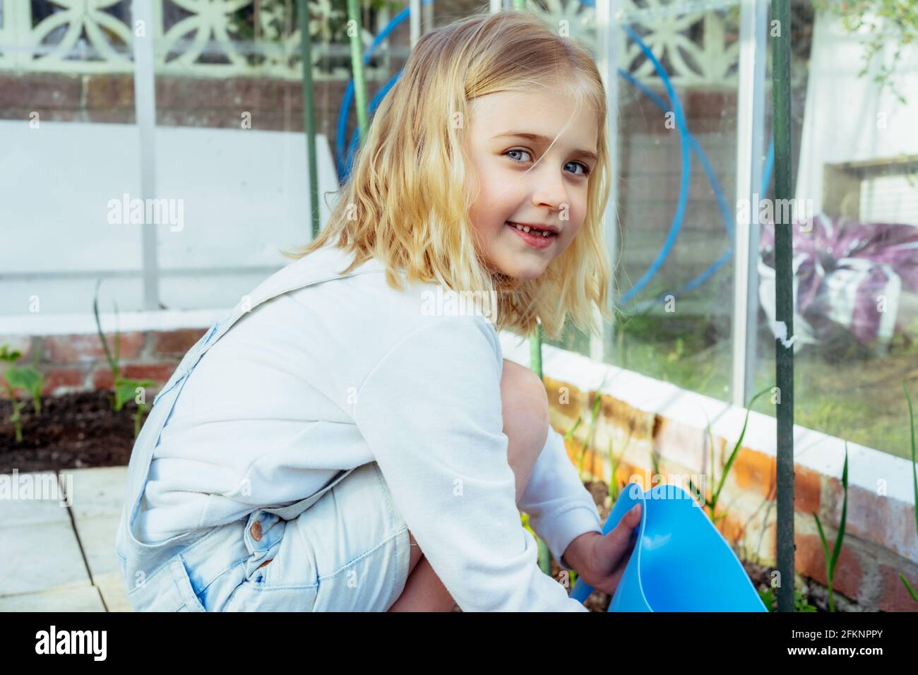 Porträt eines lächelnden zahnlosen kleinen blonden Mädchens, das Gemüse im Gewächshaus wässert. Kind hilft, sich um die Pflanzen zu kümmern. Anbauen von Lebensmitteln in der Küche g Stockfoto