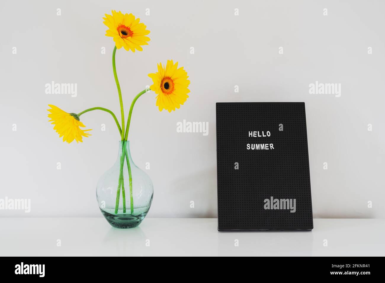 Zitat - Hallo Sommer auf dem Briefbrett und frische gelbe Gerbera Blumen in der Glasvase auf einem weißen Tisch. Sonnige Sommerstimmung Hintergrund. Minimalismus Stockfoto