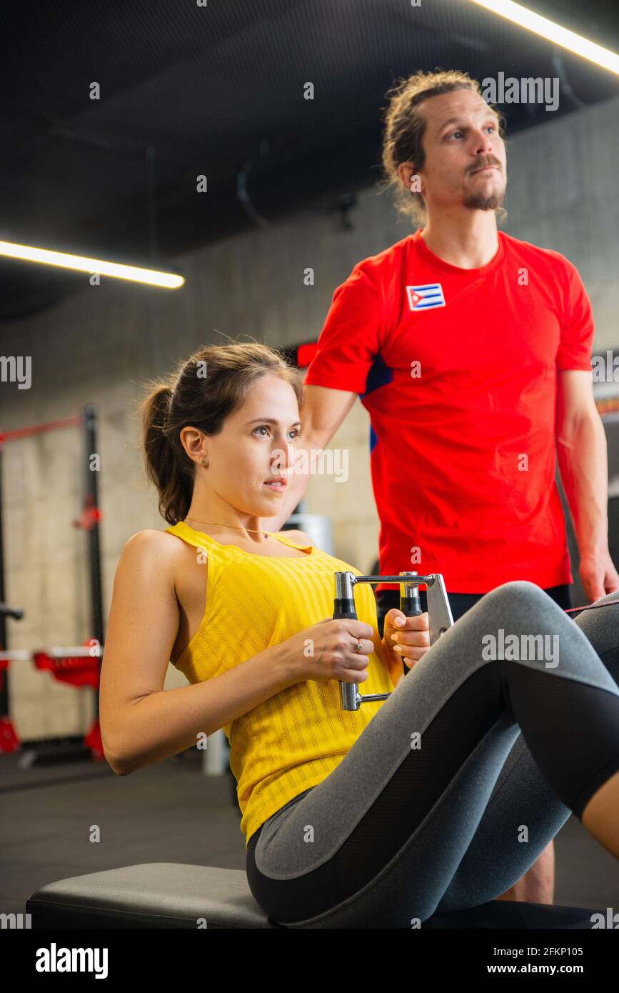 Junge kaukasische Sportlerin im modernen Fitnesscenter trainieren Auf einem Rudergerät mit persönlichem Trainer dafür sorgen, dass sie Die richtige Form beibehalten Stockfoto
