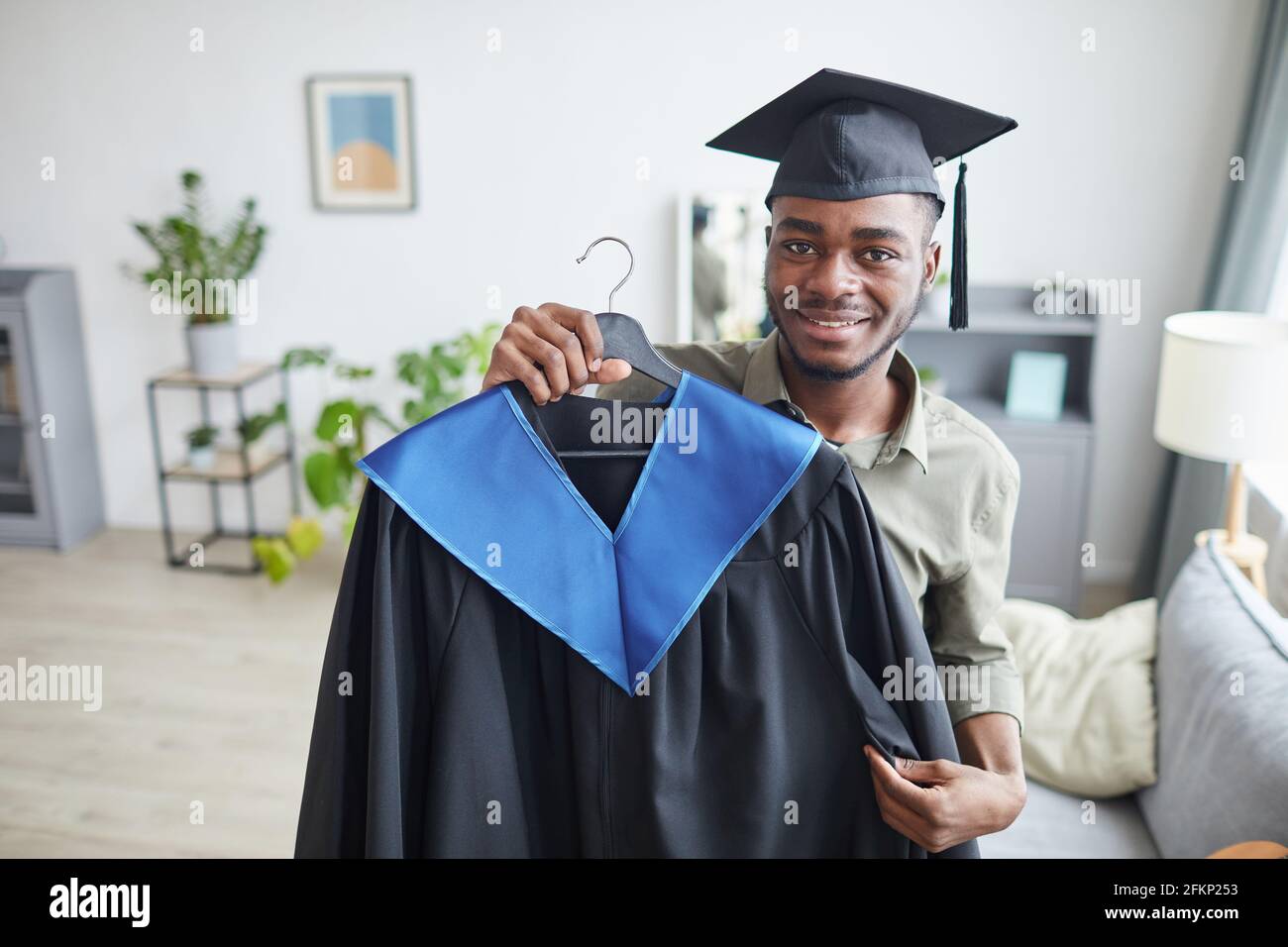 Porträt eines glücklichen afroamerikanischen Mannes mit Abschlusskleid und lächelnd an der Kamera, während er sich zu Hause auf die Zeremonie vorbereitete, Kopierraum Stockfoto