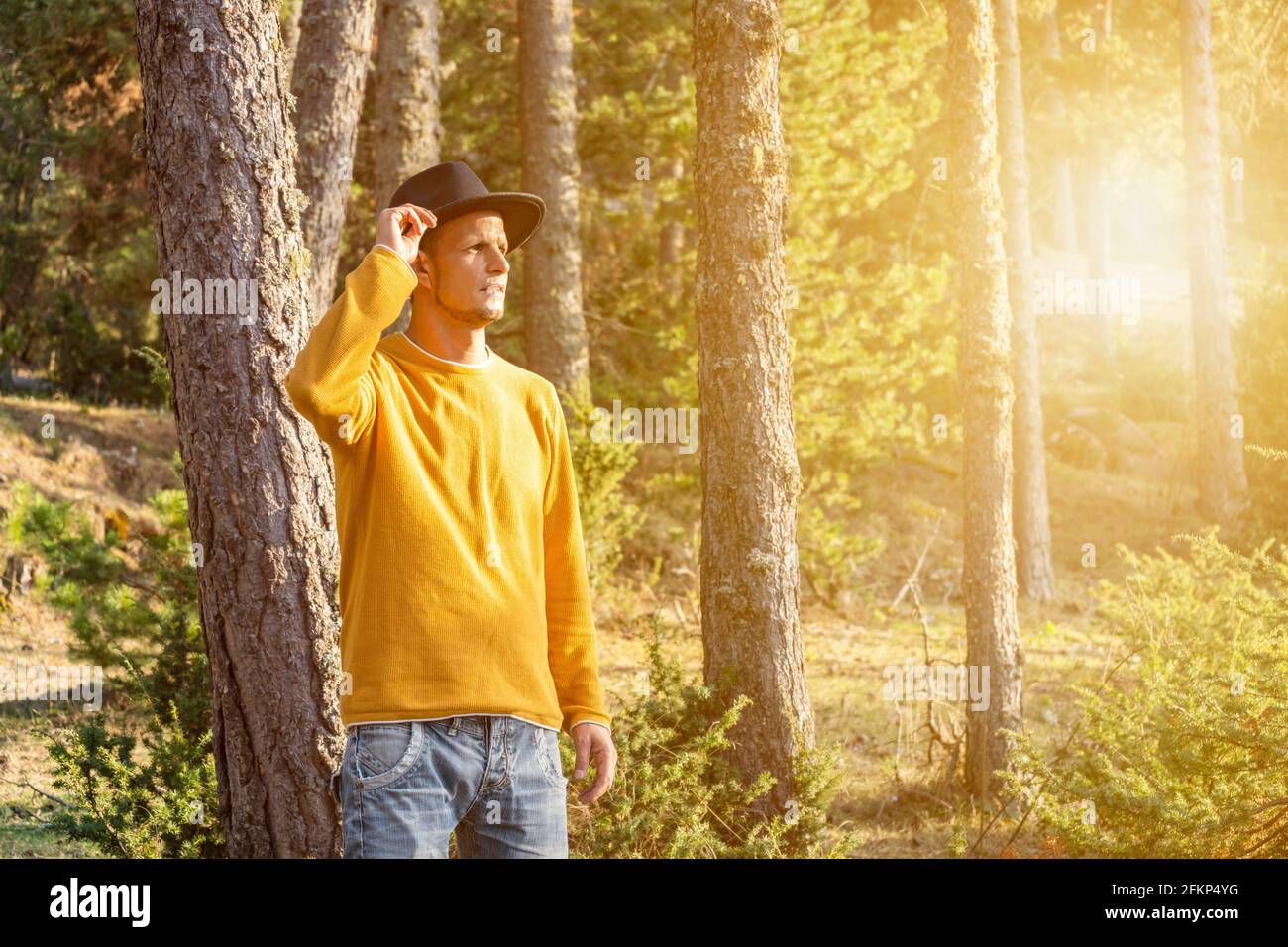 Junger stilvoller Mann im Hut im Wald.Portrait Tourist Cowboy In gelber Pullover im Wald bei Sonnenuntergang.schöne Landschaft mit Sonnenlicht und Copy Spa Stockfoto