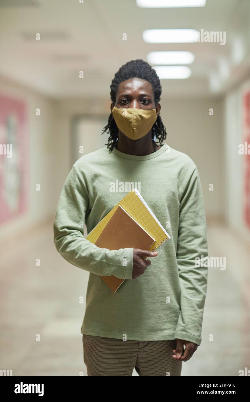 Vertikales Porträt eines jungen afroamerikanischen Mannes mit Maske in der Schule Korridor und Blick auf die Kamera Stockfoto