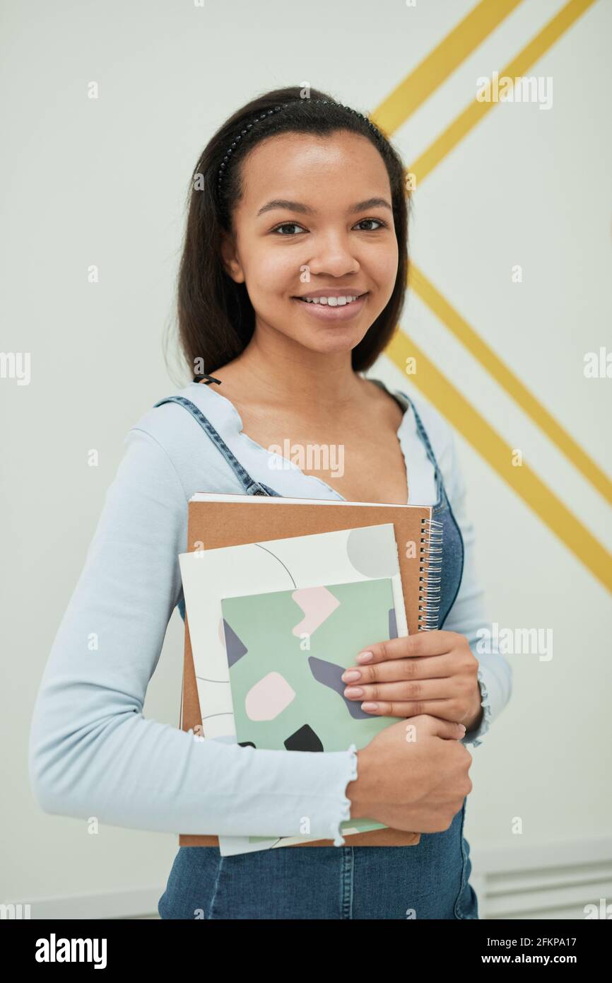Vertikales Porträt einer jungen Frau mit gemischtem Rennen, die in der Kamera lächelt Schule, während sie gegen die Wand steht und Notizbücher hält Stockfoto