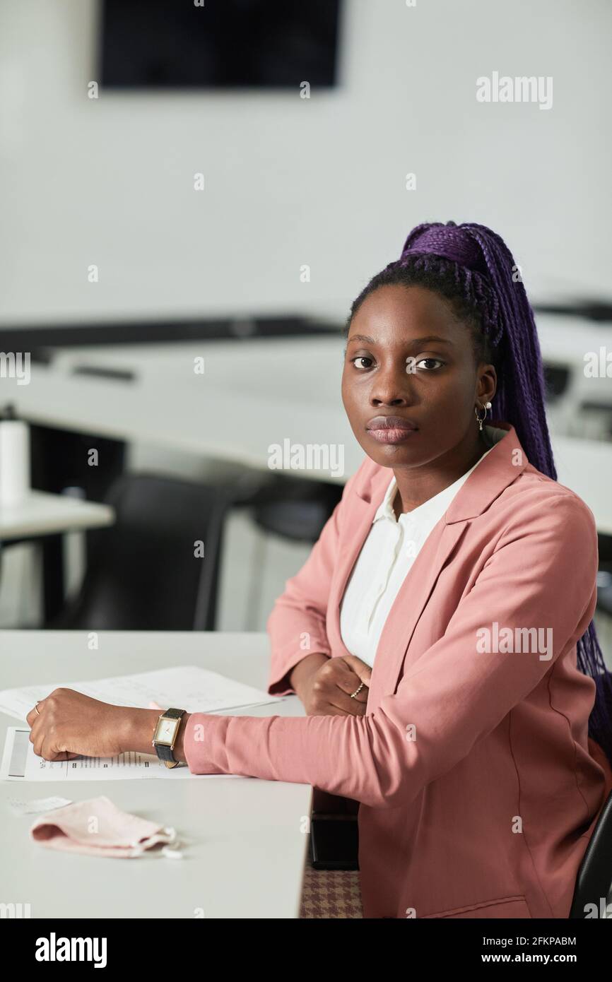 Minimales vertikales Porträt einer jungen afroamerikanischen Frau, die die Kamera anschaut In der Schule, während man im Klassenzimmer am Schreibtisch sitzt Stockfoto