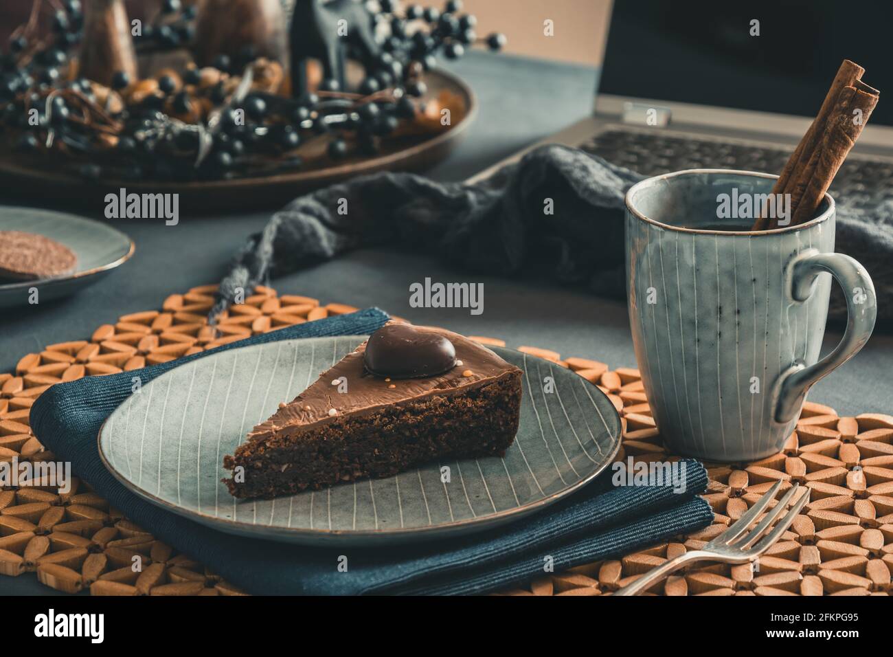 Stück hausgemachte Schokoladenkuchen und eine Tasse mit Tee und Zimtstange auf einem grauen Teller auf einem herbstlich dekorierten Tisch, Laptop im Hintergrund öffnen. Stockfoto