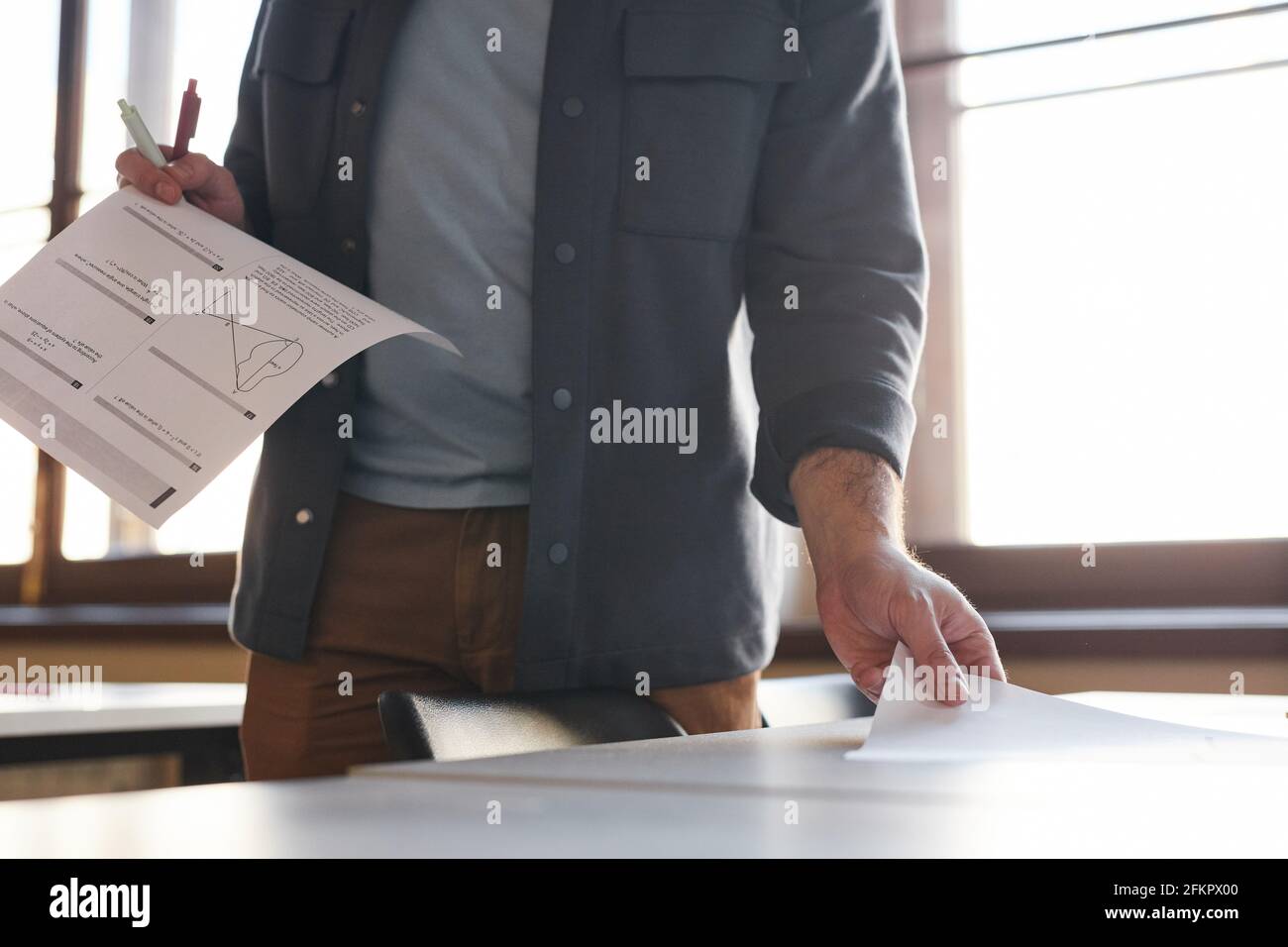 Ausgeschnittene Aufnahme eines unerkennbaren College-Professors, der Prüfungsarbeiten in einem von Sonnenlicht beleuchteten Schulsaal auslegt, Kopierraum Stockfoto