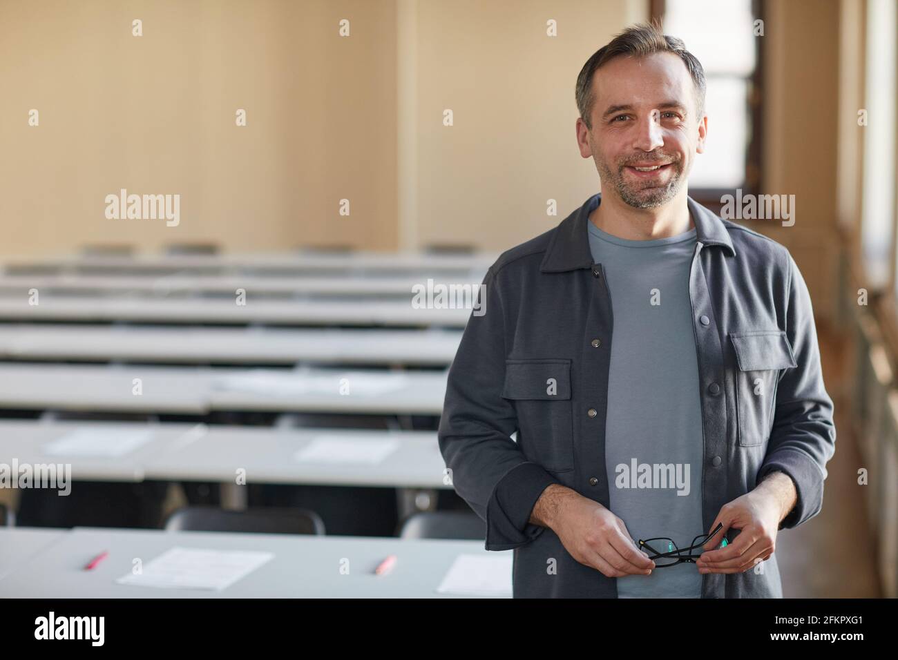 Waist-up-Porträt eines reifen College-Professors, der mit der Kamera lächelt, während er im Schulaula steht und Platz kopiert Stockfoto