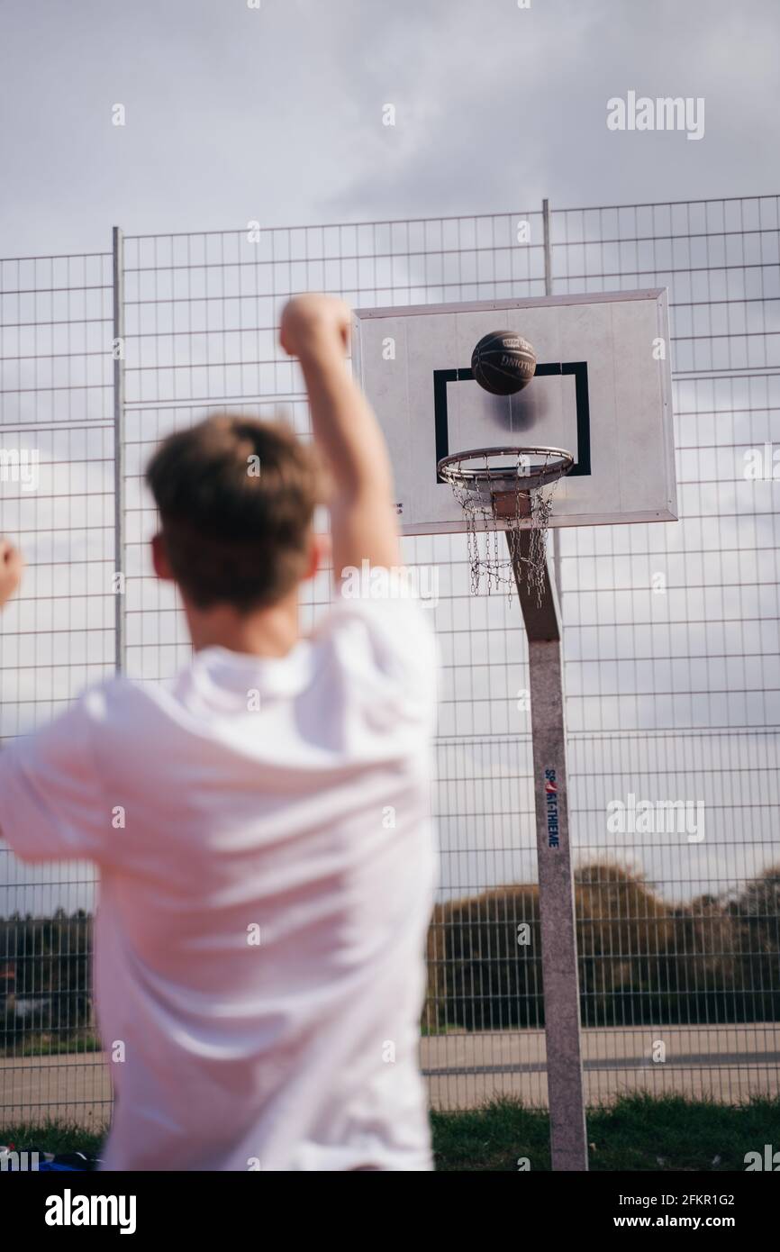 Junge/junge Mann spielt im Sommer auf einem Schulhof Basketball. Er dreht und wirft den Ball mit einem Kettennetz durch den Basketballkorb. Stockfoto