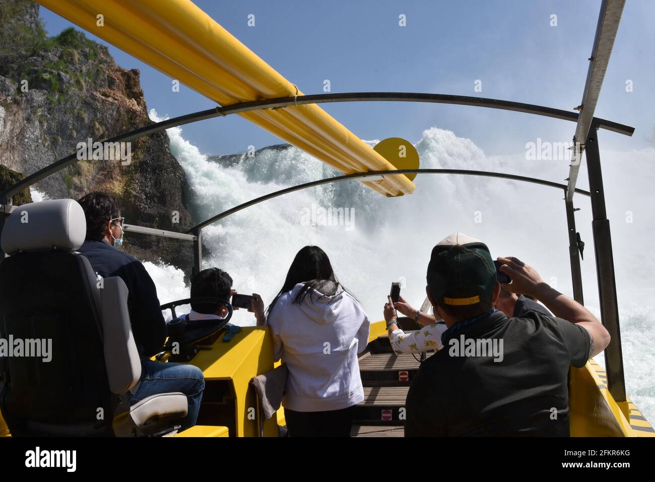 Gelbes Touristenschiff nähert sich dem Wasserfall der Rheinfälle, um das spritzende weiße Wasser zu erleben. Tourist in der Rückansicht Fotografieren Sie mit Mobiltelefonen. Stockfoto