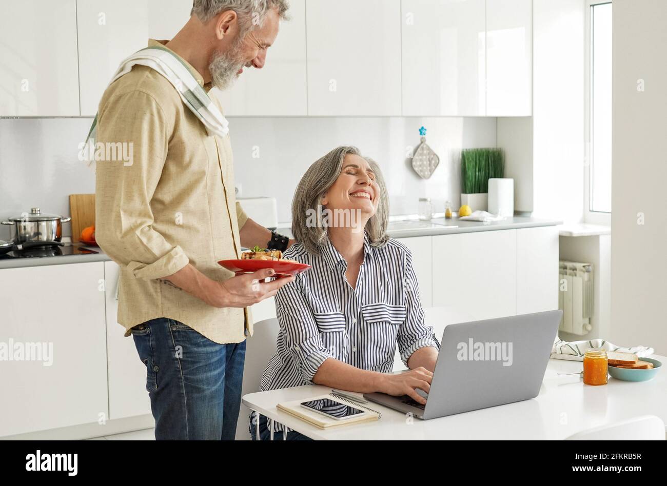 Reifer Mann bringt Frühstück zu einer glücklichen Frau, die vom Heimbüro aus arbeitet. Stockfoto