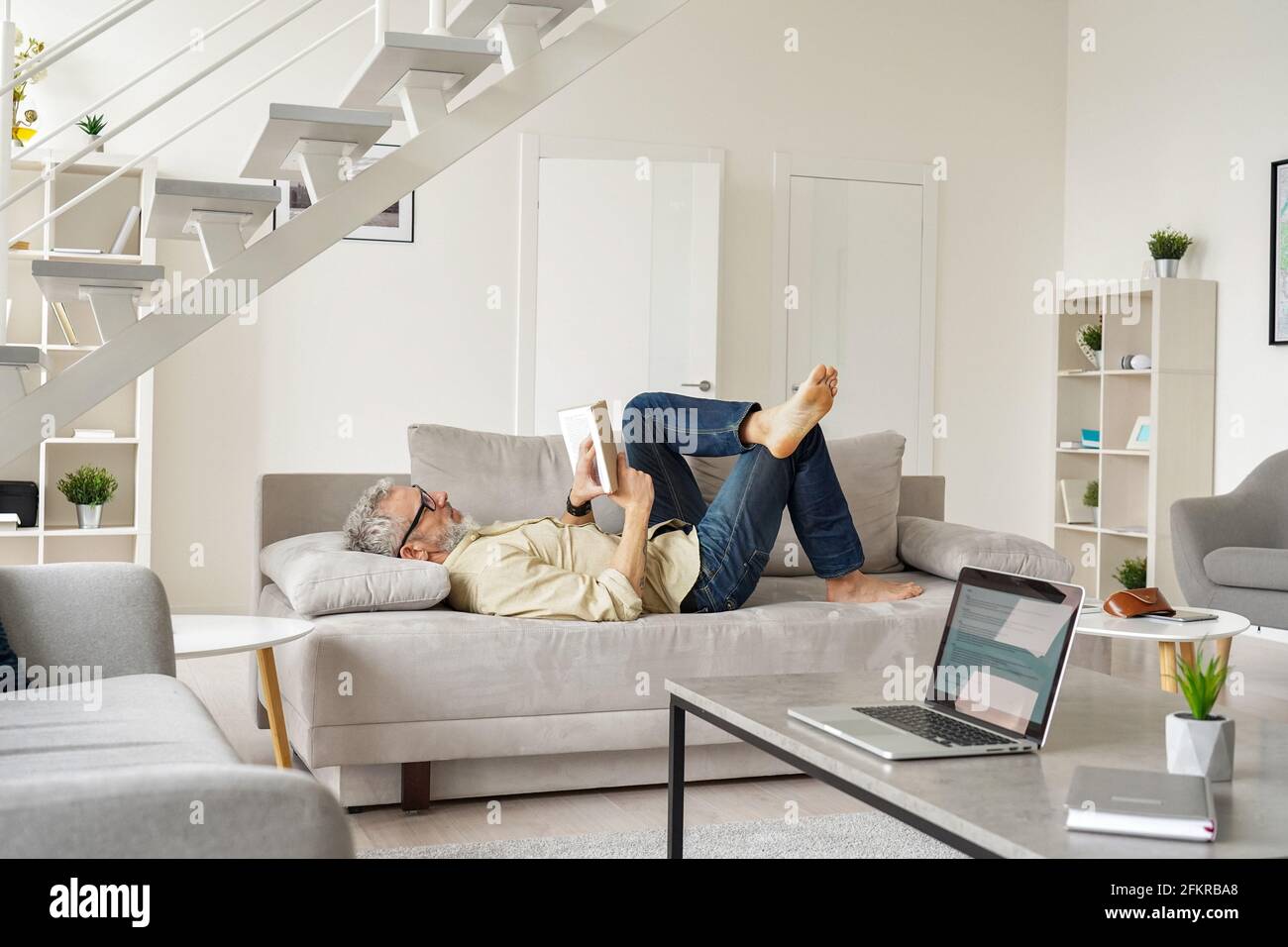 Älterer Mann mit Brille und Lesebuch beim Entspannen auf der Couch zu Hause. Stockfoto