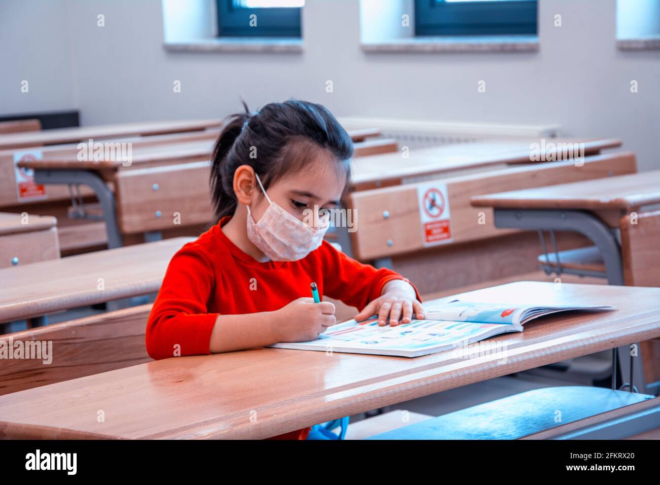 Kleines Mädchen, das im leeren Klassenzimmer mit einer schützenden Gesichtsmaske studiert Stockfoto