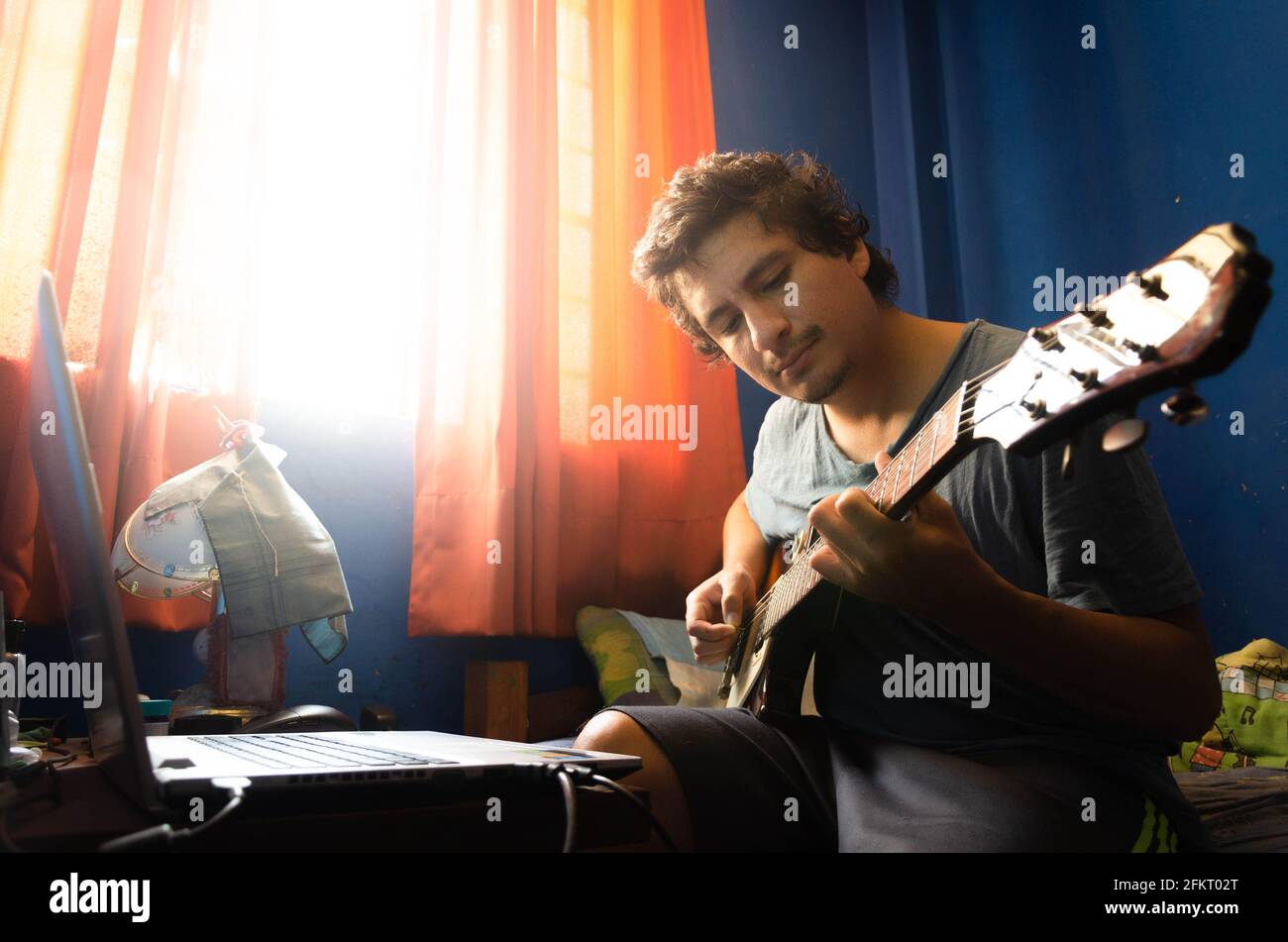 Lässiger junger Mann mit Gitarre und Laptop im Bett zu Hause. Stockfoto