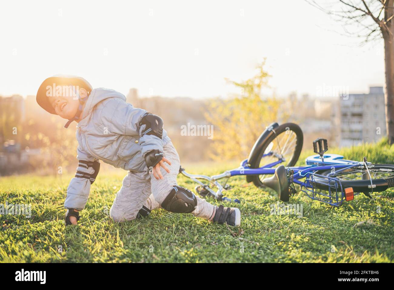 Ein Kind in einem Helm und Schutz fiel von einem Fahrrad auf das Gras und wurde nicht verletzt auf einem Sonniger Tag im Park Stockfoto