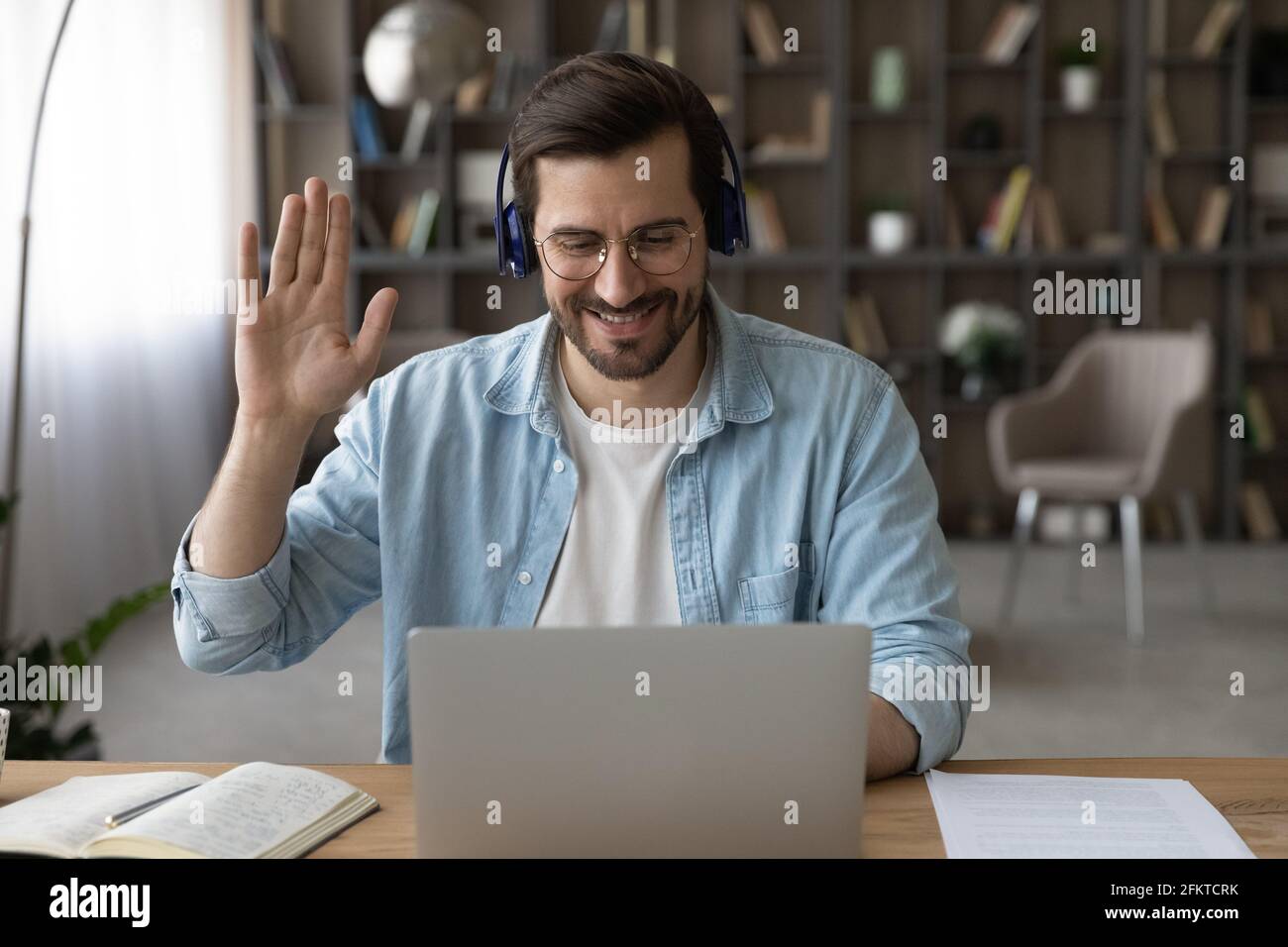 Lächelnder Mann mit Kopfhörern, der mit der Hand die Webcam anwinkt und einen Laptop benutzt Stockfoto