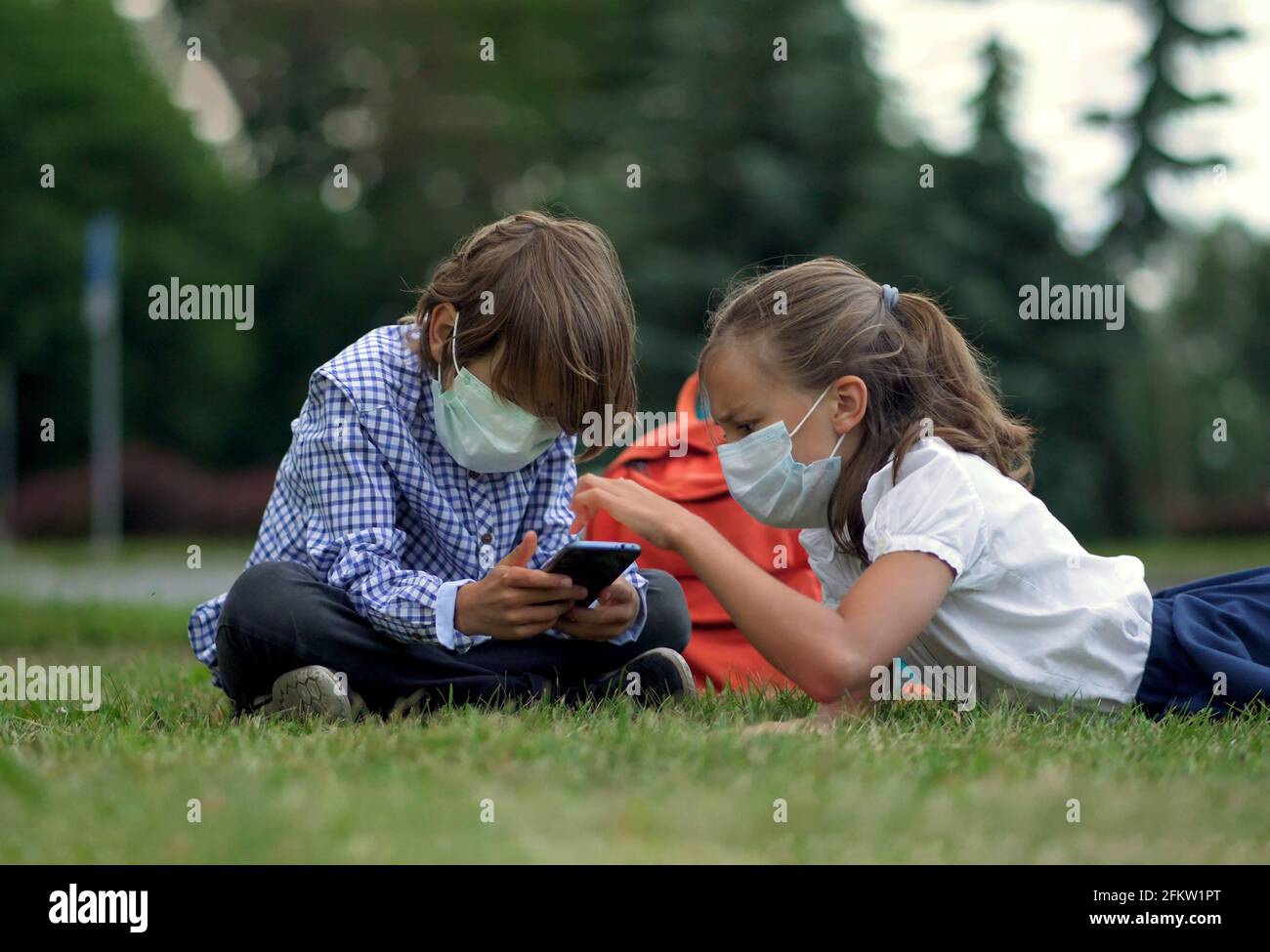 Die Kinder gehen zurück zur Schule. Niedliche Schüler mit Rucksäcken. Junge und Mädchen in Sicherheitsmasken liegen auf dem Gras Stockfoto