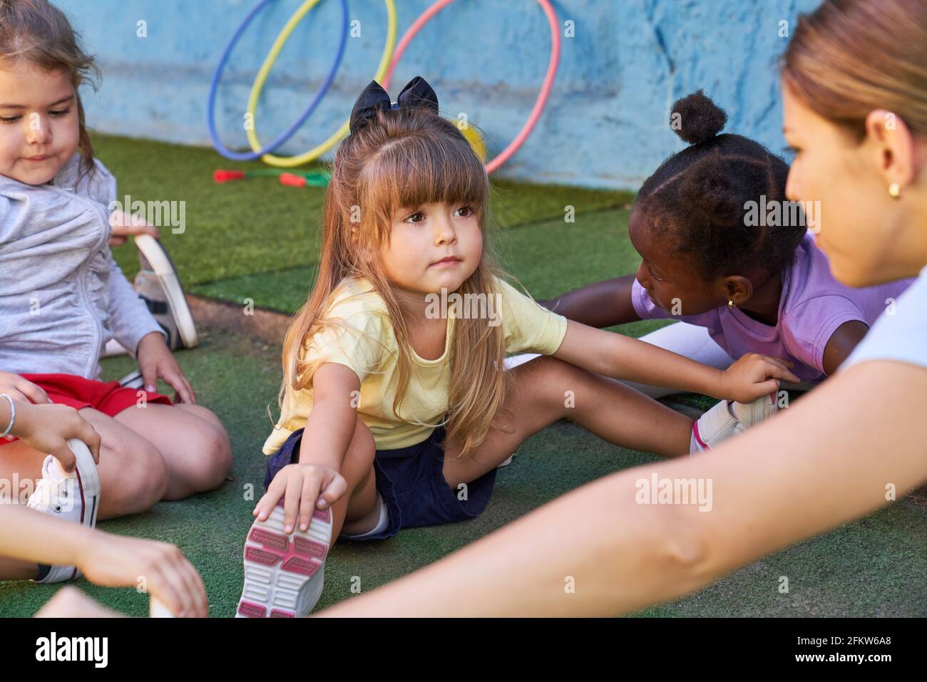 Kleines Mädchen, das sich mit anderen Kindern im Sportunterricht ausdehnt In der Kindertagesstätte Stockfoto