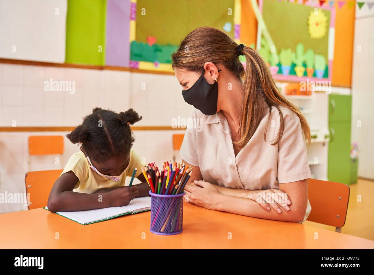 Die Kinderpflegerin kümmert sich mit einer Hausaufgabe um das afrikanische Mädchen Maske wegen Covid-19 Stockfoto