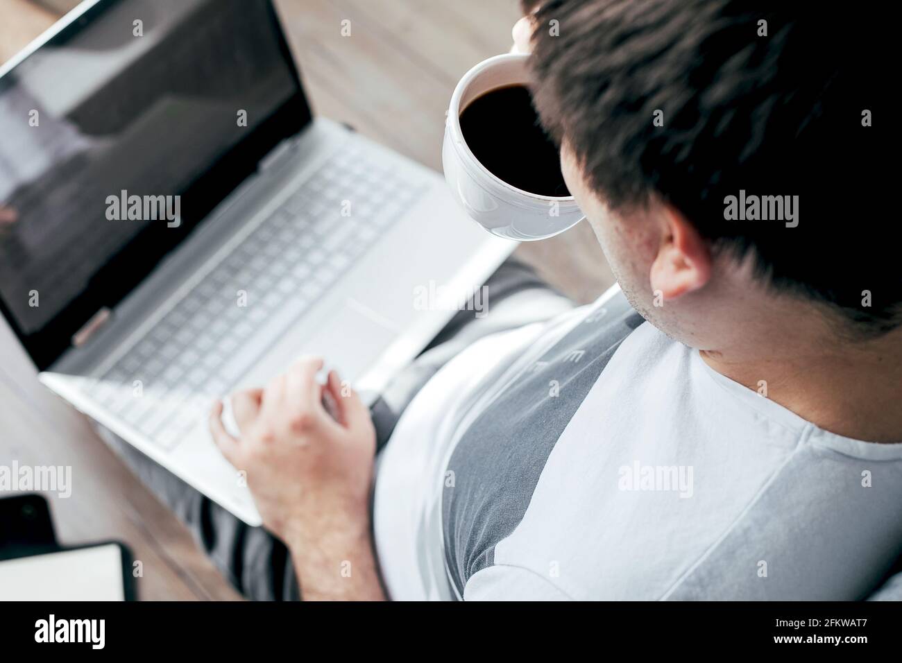 Junger Mann, der im Schlafzimmer auf dem Boden sitzt und einen Laptop benutzt und Kaffee trinkt. Arbeiten Sie von zu Hause aus. Bleib zu Hause, bleibe sicher, Quarantäne, Selbstisolierung. Top Vie Stockfoto