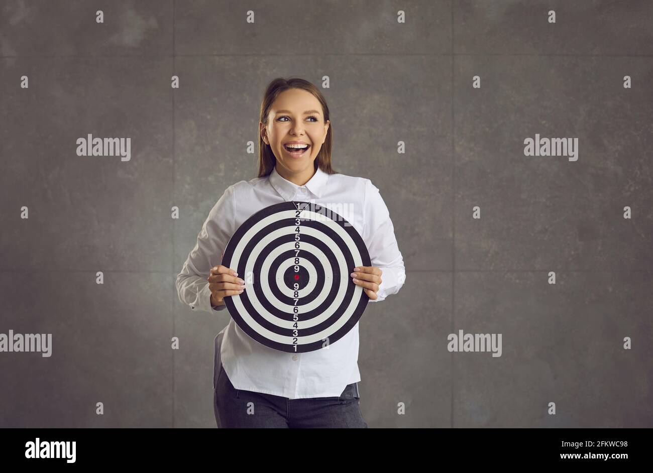 Junge, laut lachende Frau, die das Ziel der Dartscheibe im Stehen hält studiowand Stockfoto