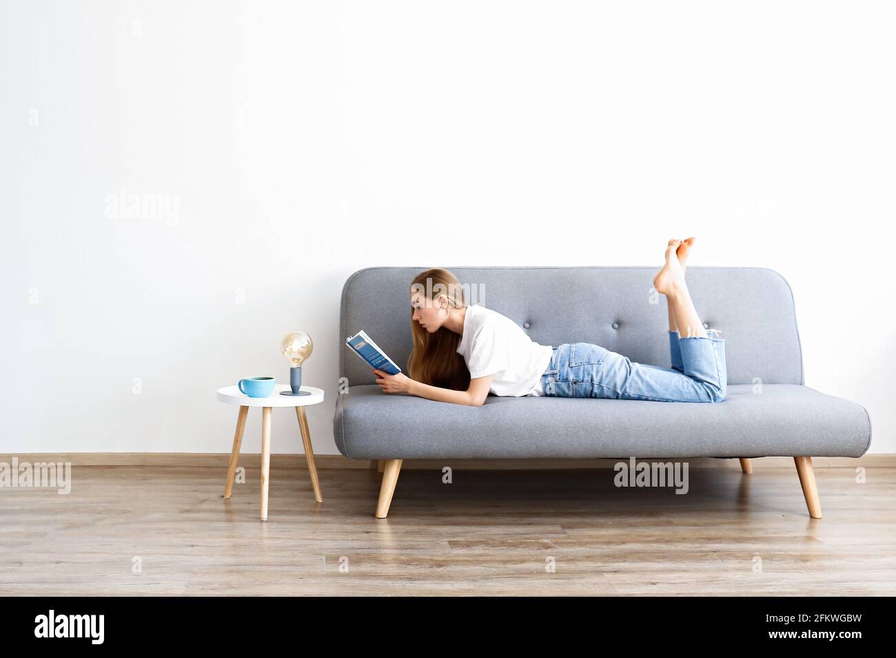Junge schöne Frau trägt weißes T-Shirt auf grauem Textil-Sofa zu Hause. Attraktive, schlanke Frau in häuslicher Situation, die sich in ihrer erhabenen Stellung auf der Couch ausruhte Stockfoto