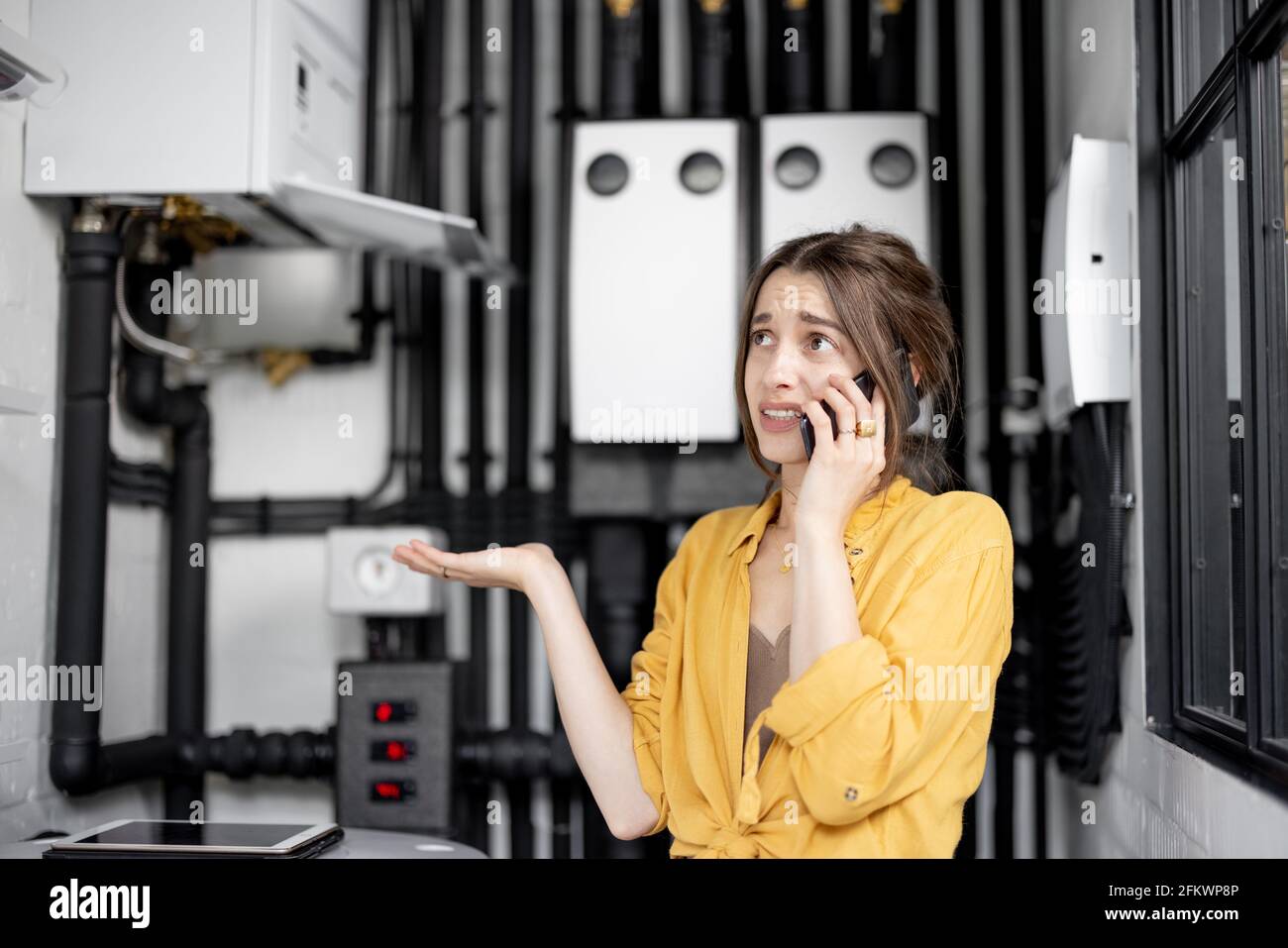 Verwirrt Frau mit Problemen mit Heizung zu Hause, spricht am Telefon versuchen, das Problem zu lösen. Konzept der weiblichen Inkompetenz in Haushaltsgeräten Stockfoto